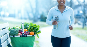 A person jogging on a bright outdoor path next to a basket filled with fresh green vegetables and colorful fruits.
